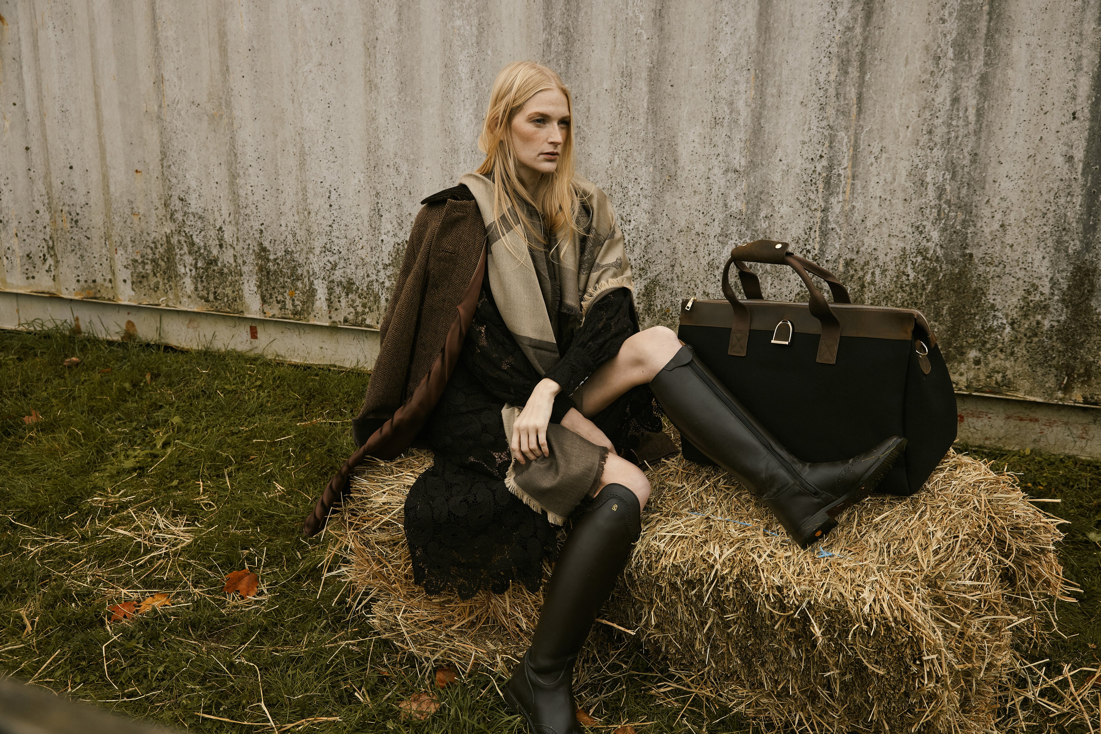 Woman sitting on a hay bale with a suitcase against a rustic wall.