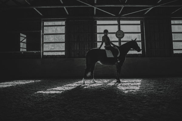 Silhouette of a person riding a horse in a dimly lit indoor arena.