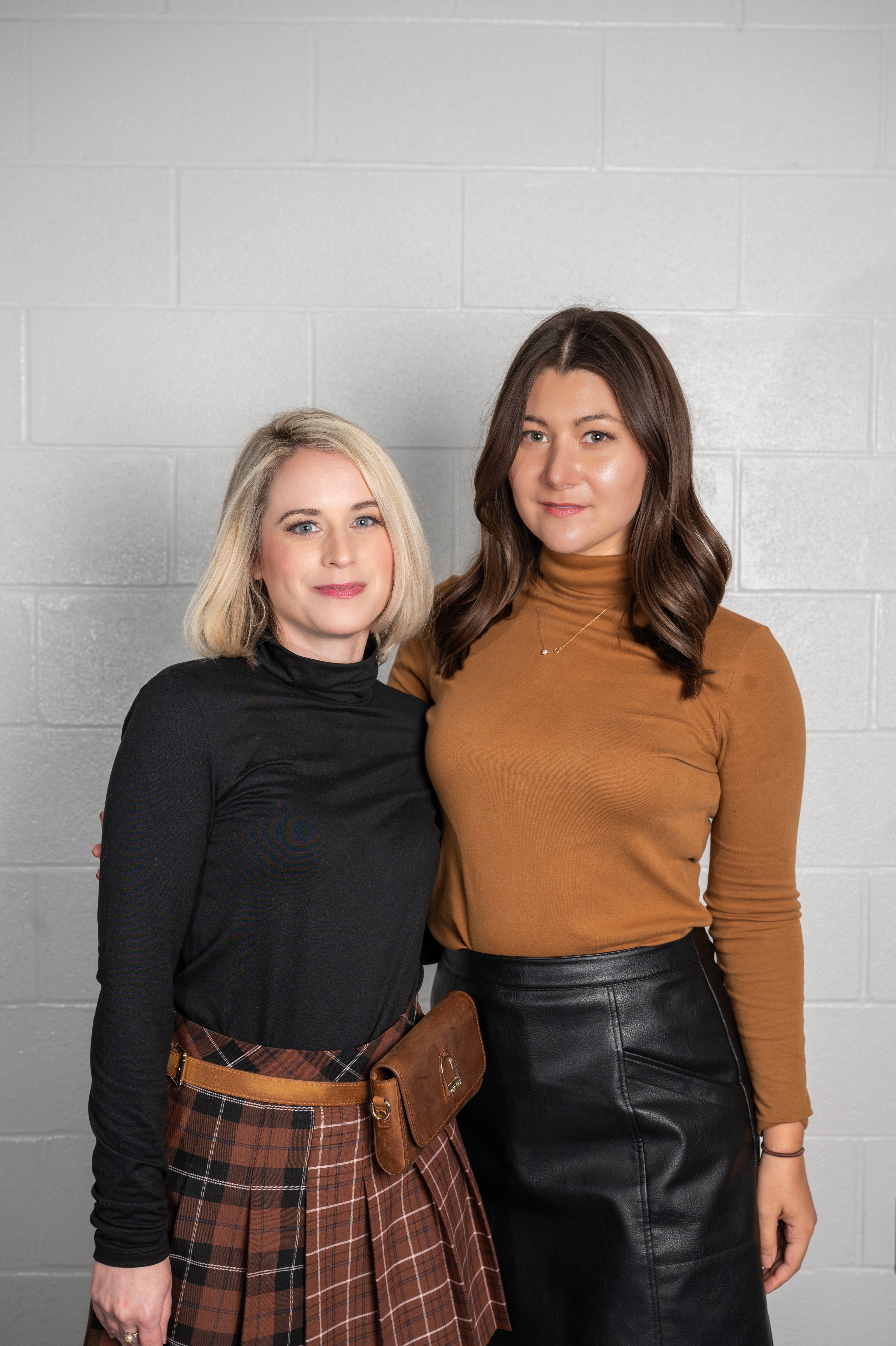 Two women standing side by side against a gray tiled wall.
