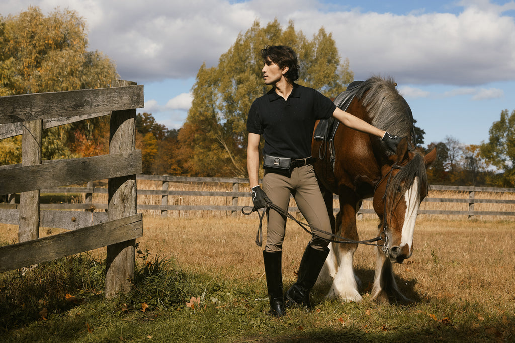 Man leading a horse through a field with a wooden fence and trees in the background