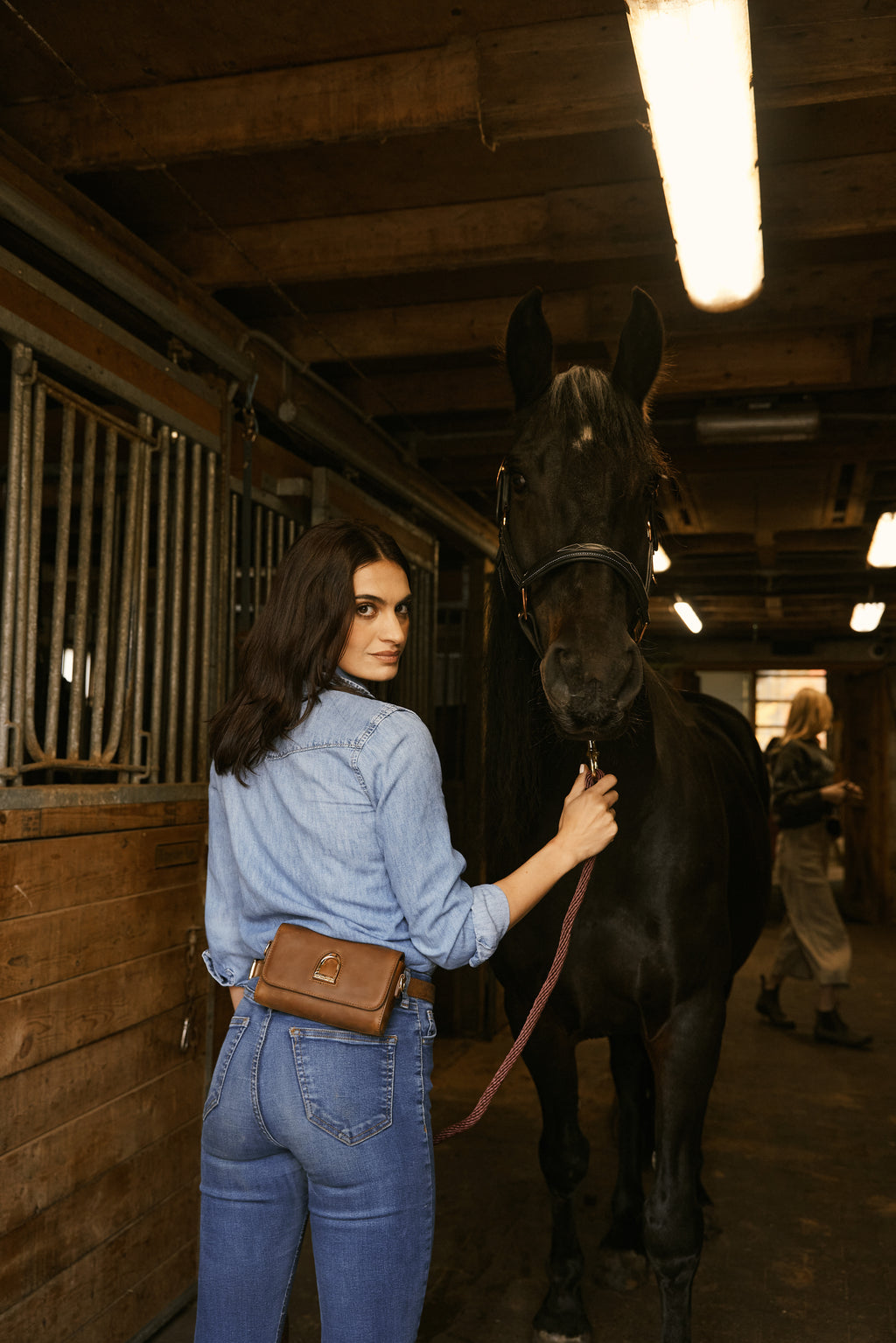 Woman in a blue denim outfit standing next to a horse in a stable.