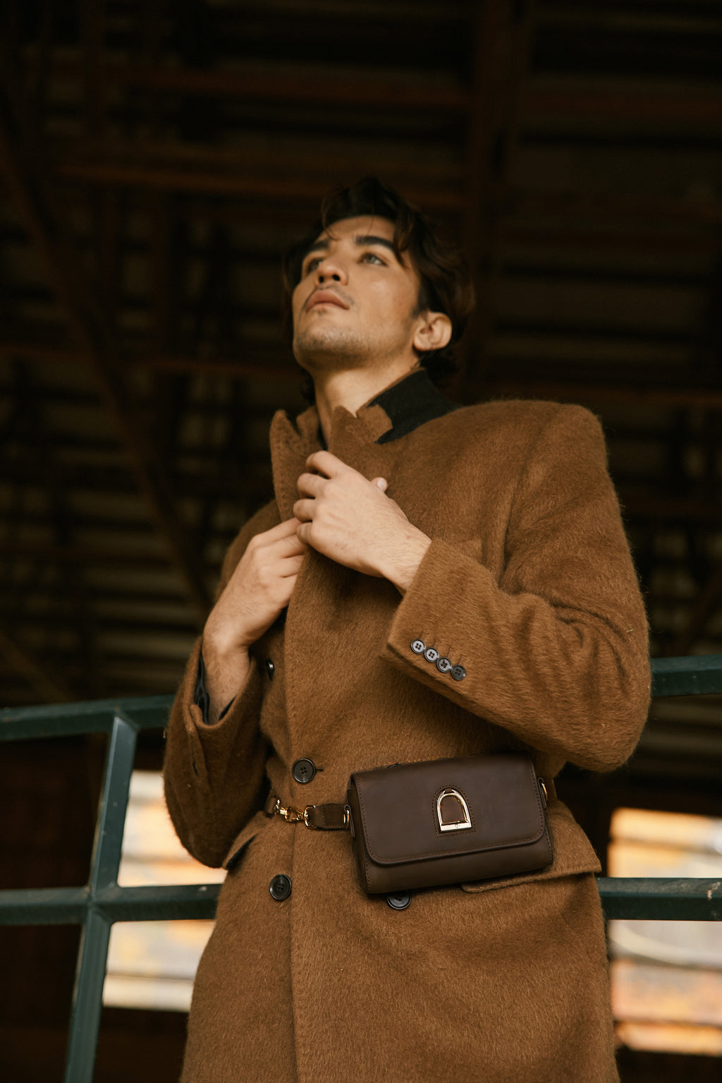 Man wearing a brown coat with a matching bag, standing against a dark background.