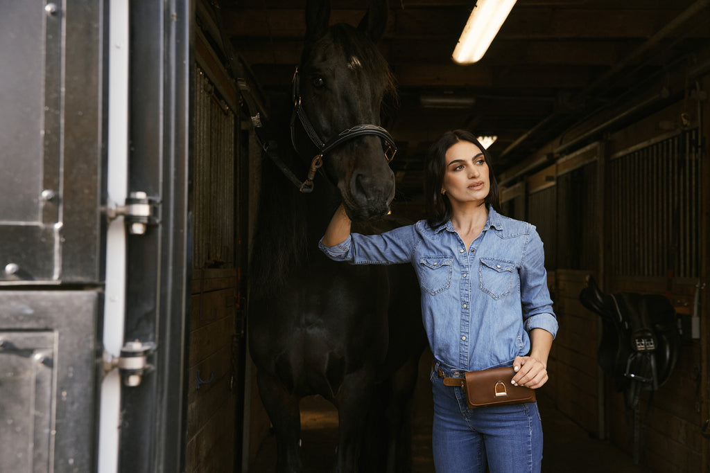 Woman standing next to a horse in a stable wearing a leather belt bag with a stirrup logo