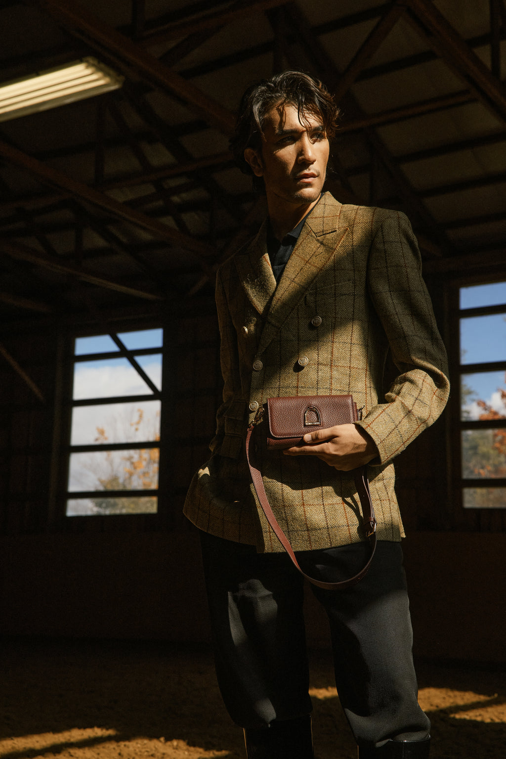 Man in a tweed jacket holding a leather bag in a dimly lit riding arena with windows.
