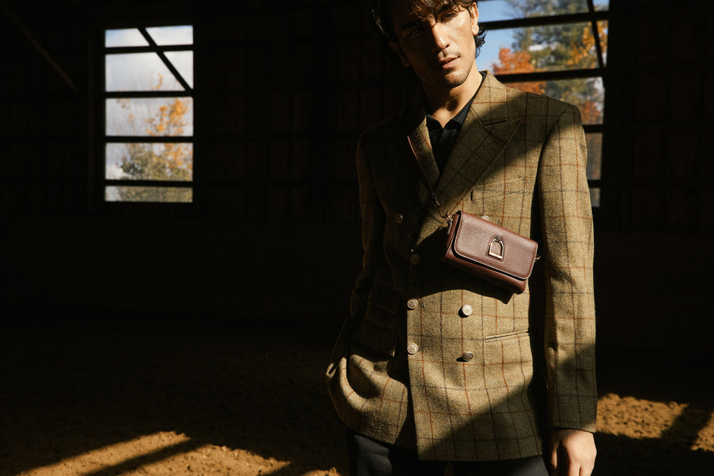 Man wearing a checkered jacket with a brown leather bag in a dimly lit room with autumn foliage outside.