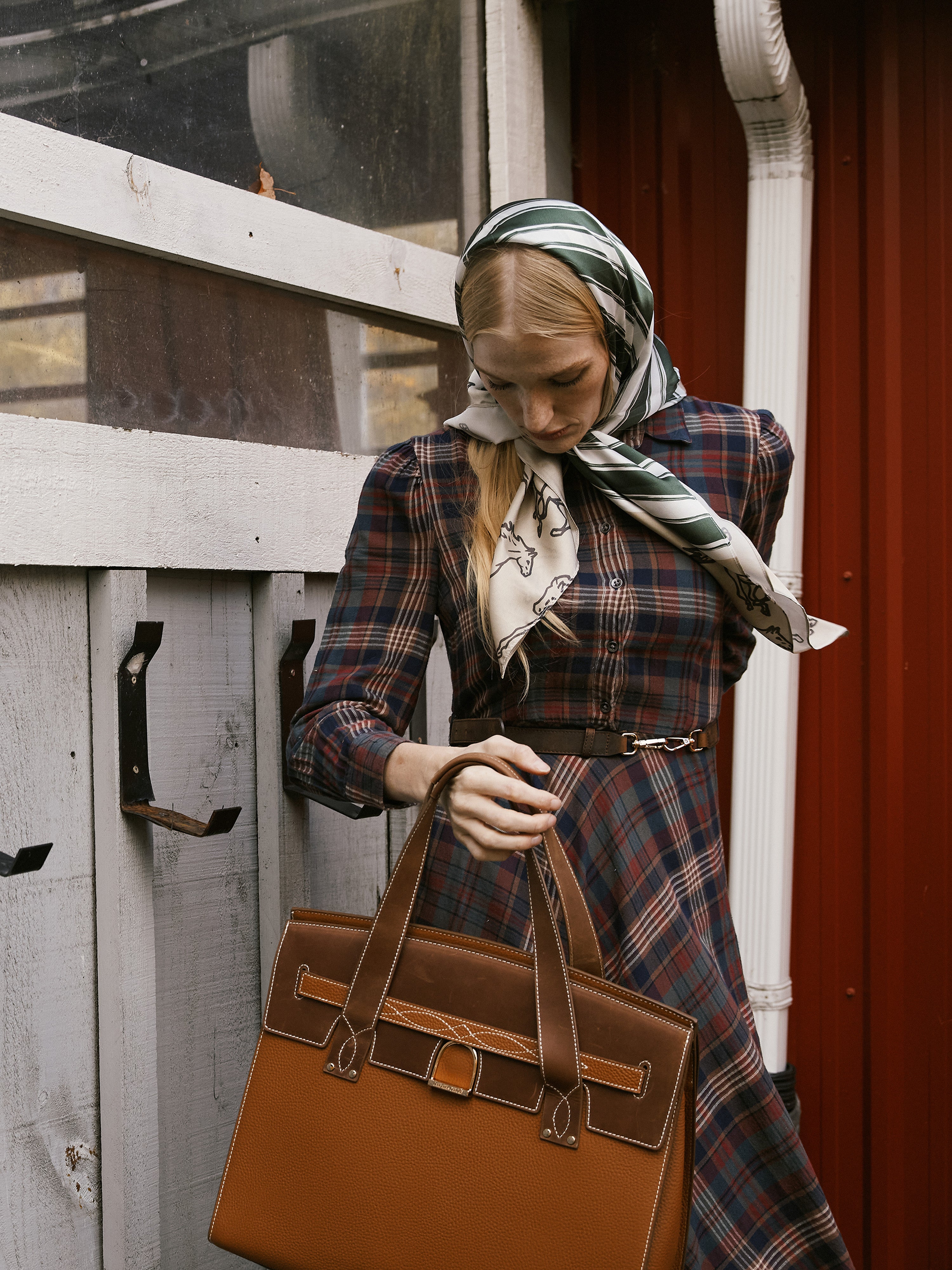 Woman in plaid dress holding a brown leather bag, standing next to a wooden structure.