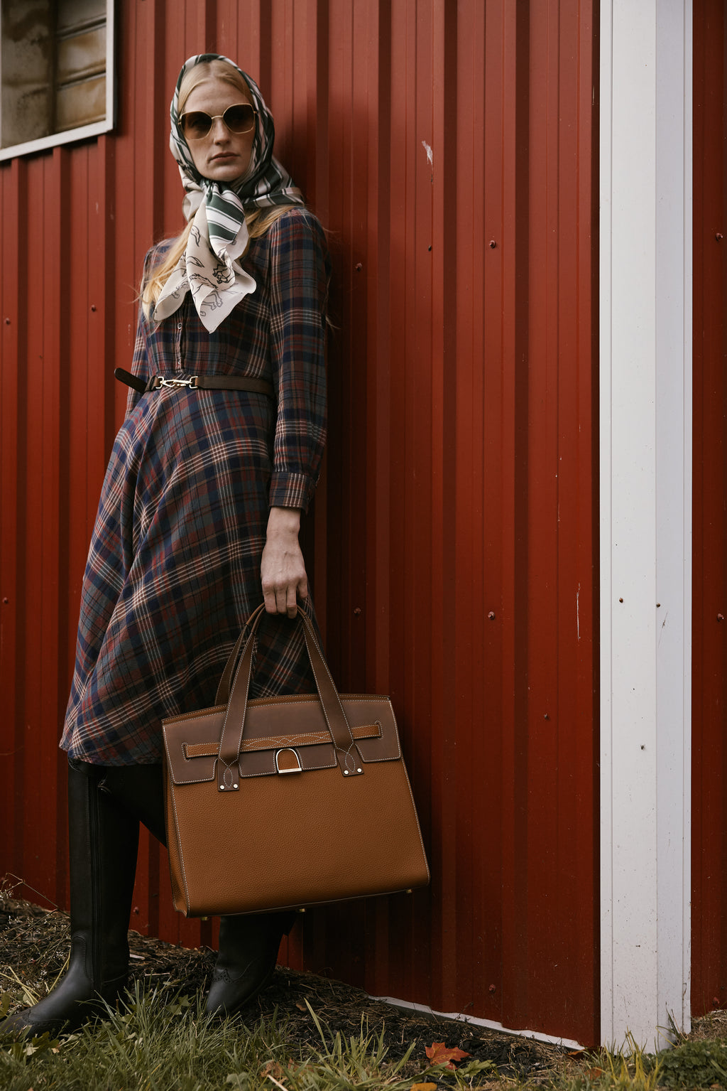Woman in plaid dress and scarf holding a brown handbag against a red barn wall.