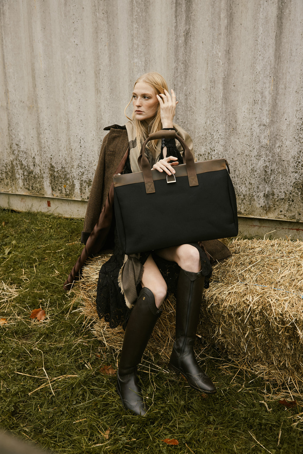 Woman holding a black leather bag sitting on hay against a rustic wall.