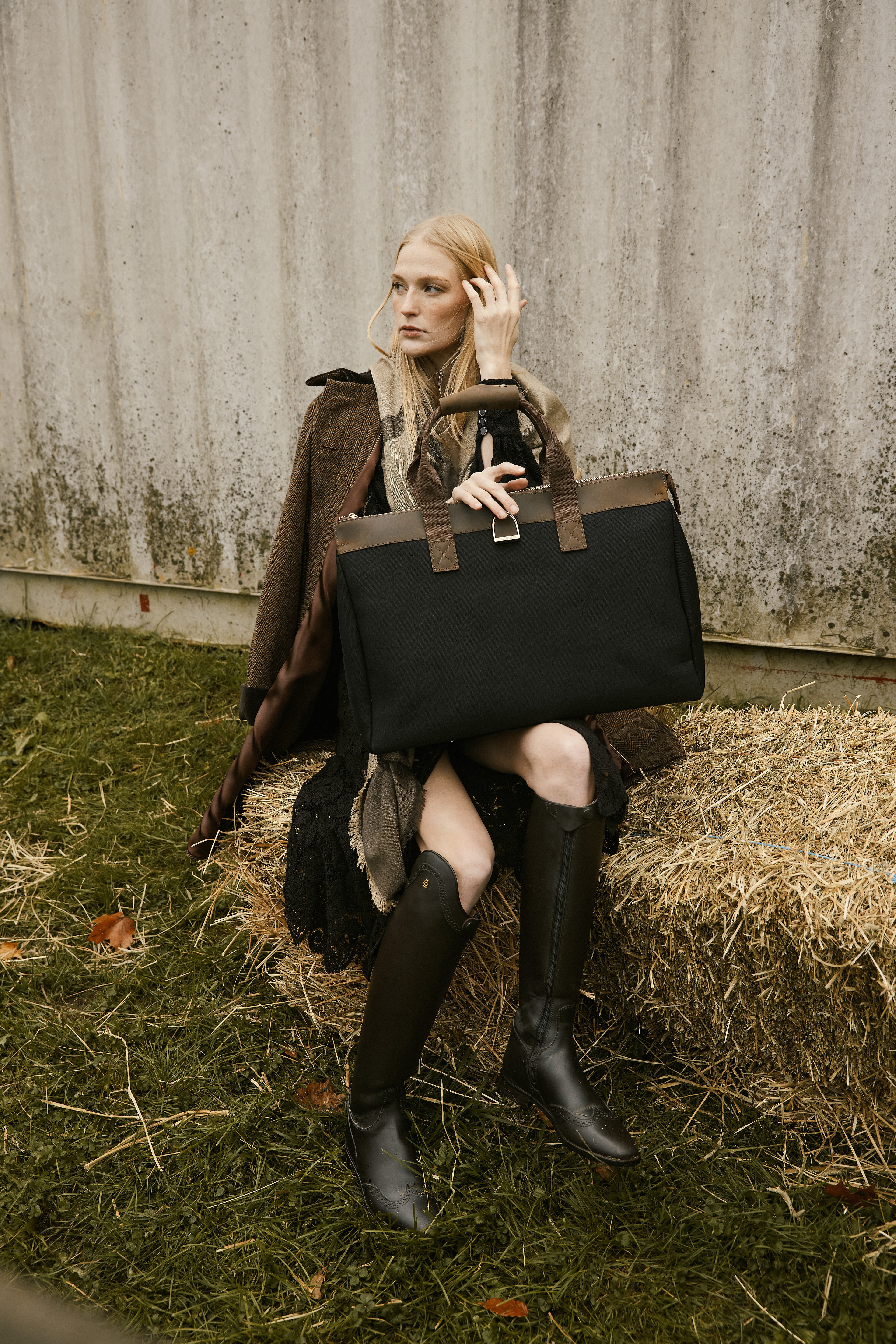 Woman holding a black leather bag sitting on hay against a rustic wall.