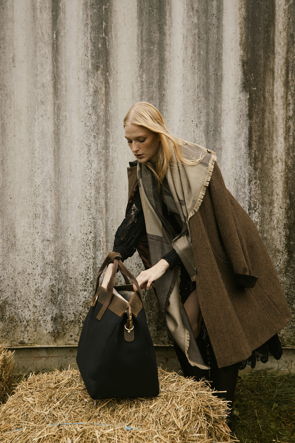 Woman in a brown coat with a patterned scarf standing next to a black bag on hay.