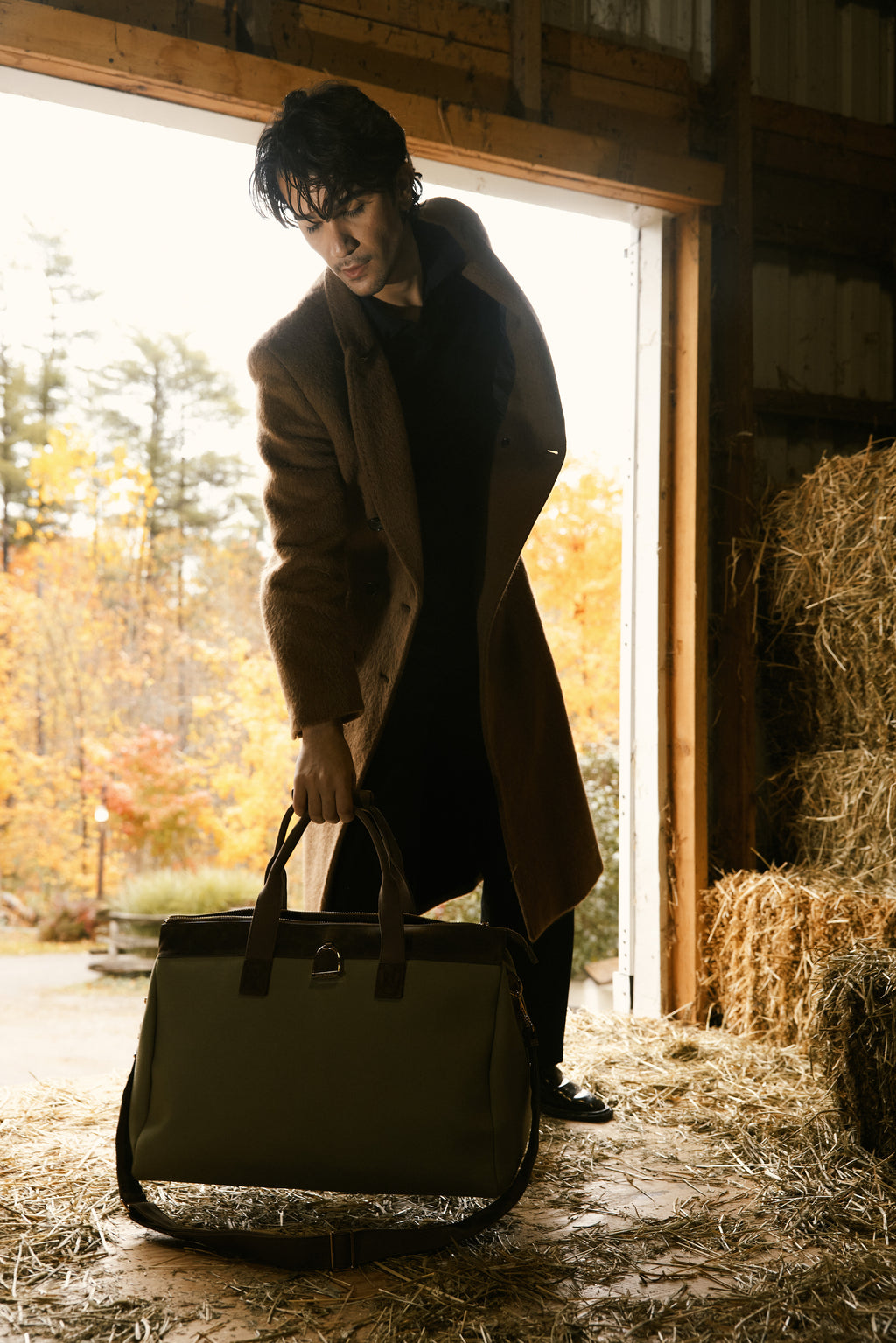 Man holding a large bag in a rustic setting with hay bales and autumn trees.