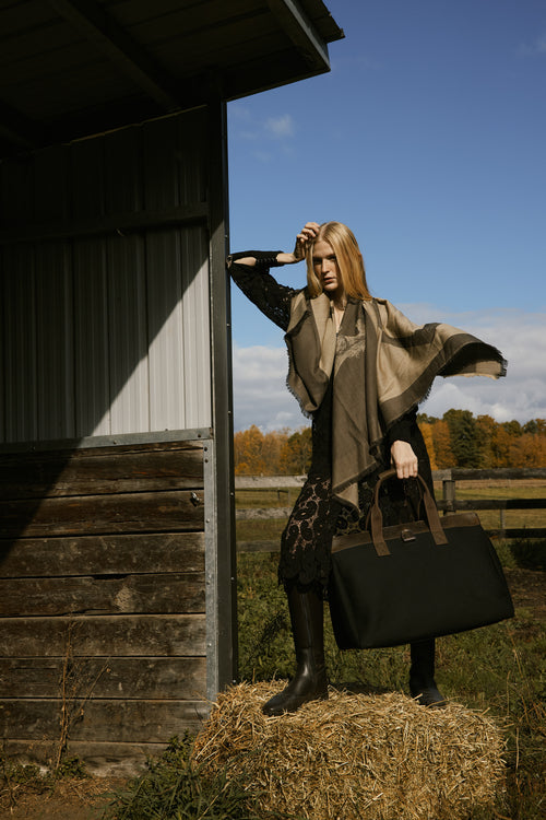 Woman standing on a hay bale holding a suitcase in a rural setting with a wooden building and blue sky.